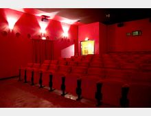 Ein kleiner Kinosaal mit roten gepolsterten Sitzen in mehreren Reihen, roten Wänden und gedämpfter Beleuchtung. Im Hintergrund befindet sich ein Ausgang mit einer beleuchteten Tür und Fenstern.