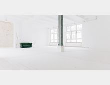 Ein heller, minimalistisch gestalteter Raum mit weißen Wänden, Holzboden und großen Fenstern, die viel Tageslicht hereinlassen. Ein grünes Chesterfield-Sofa steht an der linken Wand neben einer dekorativen Birkenstamm-Dekoration, und eine freistehende Metallstütze ist sichtbar.