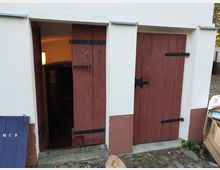 Weiße Außenwand eines Gebäudes mit zwei Holztüren in rotbrauner Farbe. Die linke Tür ist halb geöffnet und zeigt einen beleuchteten Innenbereich, während die rechte Tür geschlossen ist; davor befindet sich ein gepflasterter Boden.