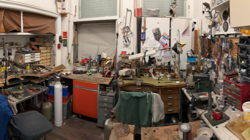 Überfüllte Werkstatt mit mehreren Arbeitstischen, Regalen und Schubladenschränken, bedeckt mit Werkzeugen, Kabeln und Kleinteilen. Im Raum sind zudem ein Waschbecken mit Küchenutensilien, Hängeschränke, Pinnwände sowie Fenster und eine Hochlagerfläche mit Stoffen und Kartons zu sehen.