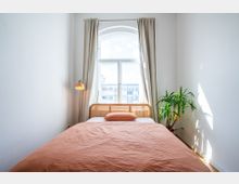 Schlafzimmer mit weiß gestrichenen Wänden und einem großen Fenster mit Rundbogen, das von hellbeigen Vorhängen umrahmt ist. Im Vordergrund steht ein Bett mit rosafarbener Bettwäsche und einem Kopfteil aus Rattan; daneben befinden sich eine Stehlampe und eine grüne Zimmerpflanze auf dem Holzboden.