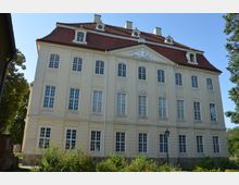 Gelbes, barockes Gebäude mit symmetrischer Fassade, zahlreichen hohen Fenstern und einem roten, geneigten Dach mit Dachgauben; im Vordergrund ein kleiner Garten mit Sträuchern und ein Laternenpfahl.