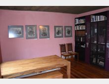 Ein kleiner Ausstellungsraum mit einer rosa Wand, an der Schwarz-Weiß-Bilder in Rahmen hängen. Im Vordergrund steht ein hölzerner Tisch, daneben sind zwei Holzstühle mit geflochtenen Sitzflächen. Rechts befinden sich Bücherregale und eine Vitrine mit Exponaten.