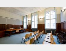 Ein Gerichtssaal mit Holzwänden und hohen, verzierten Fenstern, durch die Tageslicht fällt. Im Vordergrund befinden sich Reihen von Holzstühlen, während im Hintergrund ein langer Holztisch mit Stühlen und Lampensteht.