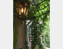 Metalllaterne mit Edison-Glühbirne hängt an einer mit Efeu bewachsenen Metallstruktur vor einem rosa Holzgebäude mit Fensterläden. Im Hintergrund ist dichter Bewuchs mit Bäumen und Sträuchern erkennbar.