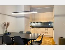 Moderne Küche mit angrenzendem Essbereich, bestehend aus einem schwarzen Tisch mit vier Stühlen. Helle Einbauküche mit weißen Schränken, grauen Wandfliesen und Holzparkettboden; eine minimalistische Hängelampe beleuchtet den Raum.