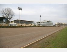 Sandbahn einer Pferderennstrecke mit angrenzenden Zuschauertribünen aus Glas und Metall sowie Flutlichtmasten; im Hintergrund moderne Gebäude.
