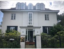 Zweistöckiges weißes Wohnhaus mit Satteldach und Dachgauben, großen Fenstern und dunklen Fensterläden. Vor dem Haus befindet sich ein kleiner Garten mit Hecken, umgeben von einem Metallzaun.