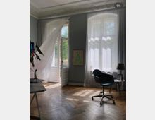 Helles Zimmer mit hohen Wänden und Stuckdecke, großen Fenstern mit weißen Vorhängen und einer geöffneten Doppeltür, die nach draußen führt. Der Raum hat einen Holzboden, einen Schreibtisch, einen schwarzen Drehstuhl und minimalistische Dekoration, einschließlich eines gerahmten Posters an der Wand.