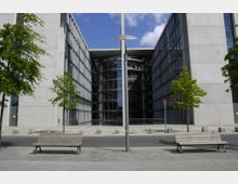 Moderne Gebäude aus Beton und Glas mit schmalem Innenhof und runden, zentralen Glasstrukturen in der Mitte. Im Vordergrund zwei Holzbänke und vereinzelte Bäume auf einem Gehweg.