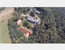 Luftaufnahme eines ländlichen Anwesens inmitten von Wald und Wiesen, bestehend aus mehreren Gebäuden mit roten Ziegeldächern und einer modernen Halle mit Solarpanelen auf dem Dach. Die Gebäude sind von Bäumen umgeben, und im Vordergrund befinden sich Rasenflächen und ein Innenhof.