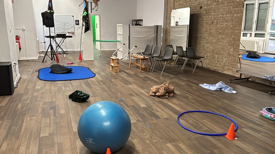 Large multipurpose studio room with wood flooring, a high ceiling with black acoustic panels and exposed pipes, and one long exposed brick wall. The space contains scattered exercise equipment such as a stability ball, cones, hoops, mats, and chairs, with windows and a bed-like platform along one side.