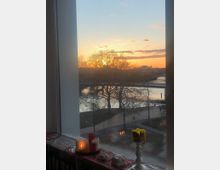 Blick durch ein großes Fenster auf eine Parklandschaft mit kahlen Bäumen und einem Fluss bei Sonnenuntergang. Auf der Fensterbank stehen brennende Kerzen und Deko-Objekte, darunter eine gelbe und eine rote Kerze.