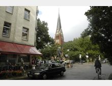 Straßenszene mit einem roten Gebäudeeckladen im Vordergrund, einem parkenden Auto, Menschen und einem Radfahrer. Im Hintergrund ragt eine Backsteinkirche mit hoher Turmspitze und Uhr aus einer Baumreihe hervor.