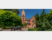 Backsteinkirche im neogotischen Stil mit spitzem Turm und Rundfenster, umgeben von Bäumen und Sträuchern. Im Vordergrund ein Zaun, ein geparkter schwarzer Geländewagen und eine Straße.