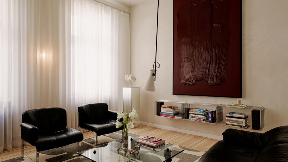 Modernes Wohnzimmer mit hohen Fenstern und bodenlangen weißen Vorhängen, hellen Holzdielen und einem gemusterten Teppich. Schwarze Ledersessel und Sofa gruppieren sich um einen gläsernen Couchtisch; an der Wand hängen ein großes dunkles Kunstbild über einem niedrigen Regal mit Büchern und es gibt eine schwenkbare Hängelampe.