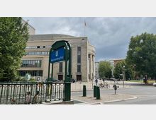 Straßenszene am Rosa-Luxemburg-Platz mit einem Bahnsteigschild der U-Bahnstation im Vordergrund. Im Hintergrund ist das Berliner Ensemble, ein markantes Theatergebäude mit säulenartiger Fassade, sowie umgebende Bäume und Gehwege zu sehen.