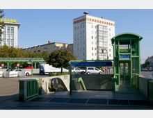 Eingangsbereich des U-Bahnhofs Innsbrucker Platz in Berlin, mit grüner verglaster Aufzugskabine und Rolltreppe. Im Hintergrund sind Wohnhäuser, ein Bürogebäude und einige parkende Autos sichtbar.