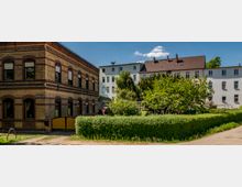 Eine Straßenansicht mit einem Backsteingebäude auf der linken Seite, das mehrere Fenster mit dunkel umrandeten Bögen besitzt, und mehreren hellen Wohnhäusern im Hintergrund. Im Vordergrund befindet sich eine grüne Hecke, daneben Bäume und ein kleiner Pfad.