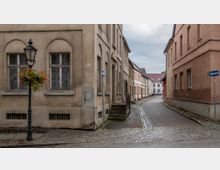 Schmale gepflasterte Straße in einer Altstadt mit beige- und orangefarbenen Gebäuden, die einfache Fensterfassungen und teils verwitterte Fassaden zeigen; links steht eine schwarze Straßenlaterne mit Blumenampel.