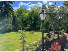 Grüne Parklandschaft mit gepflegtem Rasen, einem zentralen Brunnen mit Statue und hohen Bäumen im Hintergrund. Im Vordergrund eine schwarze Laterne, blühende Sträucher und ein kleiner Baum.