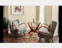 Wohnzimmer-Ecke mit zwei hellgrauen Sesseln mit geschwungenen Holzarmlehnen, einem runden Glastisch mit Holzgestell und einem abstrakten Kunstwerk an der Wand. Der Raum hat Parkettboden, beigefarbene Tapeten mit floralem Muster und ist dezent dekoriert mit Kissen und Zimmerpflanzen.