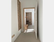 Ein schmaler, heller Flur mit weißen Wänden und sichtbaren Ziegeln in Holzrahmen; am Ende des Flurs steht ein niedriger Holzschrank mit einer Kleiderstange, daneben eine grüne Zimmerpflanze.