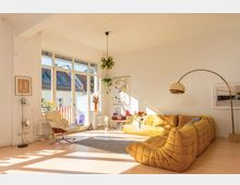 Helles Wohnzimmer mit großen Fenstern, die viel Tageslicht einlassen. Der Raum ist modern eingerichtet, mit gelben Sofa-Elementen, einem geschwungenen Stehlampenbogen, Holzboden, Zimmerpflanzen und Dekorationen, sowie einer weißen Wand im Hintergrund.