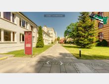 Ein gepflasterter Weg führt durch einen begrünten Innenhof mit modernen und historischen Gebäuden, darunter ein rotes Backsteingebäude und ein hell verputztes Haus. Im Vordergrund steht eine rote Infotafel mit der Aufschrift „GLS Campus Berlin“.