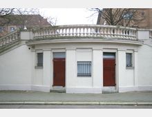 Gebäudefront eines öffentlichen Toilettenhäuschens aus hellem Putz mit rotem Holztor und kleinen vergitterten Fenstern. Darüber verläuft eine weiße, verzierte Balustrade, im Hintergrund sind Bäume und ein mehrstöckiges Gebäude zu erkennen.