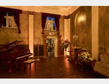 Eleganter, klassisch eingerichteter Raum mit einem polierten Flügel, Kerzenleuchtern und Blumenarrangements. Die Wände haben eine historische Optik, dekoriert mit dunkelroten Vorhängen, während eine geöffnete Tür mit Ornamentrahmung den Blick in einen weiteren Raum freigibt.