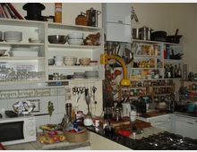 Eine voll ausgestattete Küche mit offenen Regalen, die Geschirr, Töpfe, Gläser, Gewürze und andere Kochutensilien enthalten. Im Vordergrund befinden sich ein Gasherd und eine Arbeitsfläche mit verschiedenen Küchengeräten.