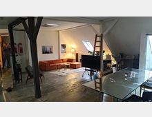 Wohnraum in einem Dachgeschoss mit offener Gestaltung, sichtbaren Holzbalken und schrägen Wänden. Im Vordergrund ein Glastisch mit Stühlen, dahinter eine orangefarbene Eckcouch, ein Flachbildfernseher und eine Leiter, die an einer Wand lehnt; Tageslicht fällt durch ein Dachfenster ein.