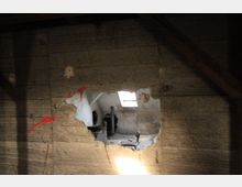 Ansicht durch ein großes Loch in einer beschädigten Wand aus Beton oder Zement. Dahinter ist ein Raum mit einem Schreibtischstuhl, einem Dachfenster und Sicht auf rohe Wände und ein freiliegendes Rohr zu erkennen.