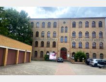 Backstein-Gebäude im Industriestil mit mehreren Stockwerken und rot umrandeten Fenstern, erkennbar beschriftet mit "Drahtgeflecht-Gitter" und "Drahtzaun-Fabrik". Im Vordergrund ein gepflasterter Hof mit geparkten Autos und einer angrenzenden Garage.