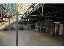 Innenansicht einer alten Industriehalle mit hohen Decken, sichtbaren Stahlträgern und einem Oberlichtdach. Die Wände bestehen aus verwittertem Mauerwerk, der Betonboden ist glatt, und moderne Beleuchtung ist an der Stahlkonstruktion montiert.