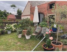 Hinterhof mit einer Ziegelsteinfassade eines alten Gebäudes, Gartenmöbeln und zahlreichen Pflanzen in Töpfen, darunter Rosen, Olivenbäume und Hortensien. Im Hintergrund sind weitere Gebäude mit roten Ziegeldächern sowie eine Steinmauer sichtbar.