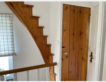 Treppenhaus-Ecke in einem Wohnhaus mit einer hölzernen, geschwungenen Treppe, einer naturbelassenen Holztür mit Metallgriff und einem Fenster mit kariertem Rollo. Im unteren Bereich ist ein Holzgeländer mit weißen Streben sichtbar.