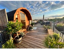Holzterrasse auf einem Dach mit Blick auf die Stadt, eingerichtet mit Pflanzen in Töpfen, Gartenmöbeln, einem Grill und einer kleinen Holzsauna mit gewölbtem Dach; im Hintergrund ist ein Fernsehturm sichtbar.