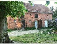 Backsteinbauernhaus mit rotem Ziegeldach und kleinen Fenstern, flankiert von einer Wiese und blühenden Fliederbüschen. Im Vordergrund steht ein großer Baum, dessen grüne Blätter einen Teil des Gebäudes überdecken.