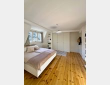 Helles Schlafzimmer mit Holzdielenboden, weißem Bett und großen Fenstern mit Vorhängen an der linken Seite. Gegenüber stehen eingebaute Kleiderschränke mit mehreren Türen; in der Ecke befindet sich ein schmales Regal und eine Deckenlampe hängt mittig.