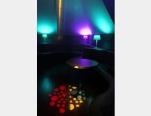 Dunkler Lounge- oder Clubbereich mit halbrunden Polsterbänken entlang der Wände und einem runden Tisch in der Mitte. Bunte Akzentbeleuchtung in Türkis und Violett sowie kleine Tischlampen und Lichtpunkte auf dem Holzboden prägen die Szene.