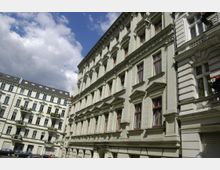 Mehrstöckige Gründerzeitgebäude in einer städtischen Straßenszene, gekennzeichnet durch verzierten Fassadenschmuck, hohe Fenster mit Holzrahmen und ein helles Putzdekor. Im Hintergrund blauer Himmel mit weißen Wolken.