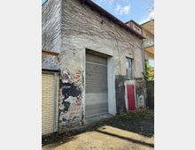 Außenansicht eines älteren Nebengebäudes mit verwitterter, teils freiliegender Ziegel- und Putzfassade, überwachsen von kahlen Kletterranken. In der Front befinden sich ein großes graues Rolltor, ein Fenster und eine kleine rote Tür; davor liegt ein gepflasterter Hof zwischen angrenzenden Wohngebäuden.