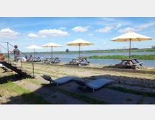 Flussufer mit Picknickbereich, bestehend aus Holzbänken und Tischen unter großen, hellen Sonnenschirmen; im Vordergrund befinden sich Liegestühle und eine Person auf einer Treppe. Im Hintergrund ist ein breiter Fluss mit grasbewachsenen Ufern und einem bewölkten Himmel zu sehen.