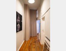 Schmaler Flur mit hellen Wänden und einem Holzboden. Am Ende des Flurs hängt ein beiger Vorhang, daneben ist eine teilweise geöffnete weiße Tür sichtbar. Links hängt ein modernes Kunstbild, rechts lehnt ein gerahmtes Bild an der Wand.