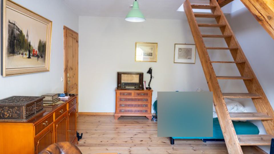 Wohnraum mit Holzfußboden, Kommode und gerahmten Bildern an hellen Wänden, beleuchtet von einer grünen Deckenlampe. Rechts führt eine offene Holztreppe zu einer Empore, darunter steht ein Bett oder Sofa.