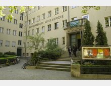 Fassade des Gemeindehauses Luisenkirche mit beige gestrichenem Gebäude, zahlreichen Fenstern und einem Eingang mit Treppe. Rechts vom Eingang stehen zwei Personen, und ein Schaukasten mit farbigen Aushängen ist im Vordergrund zu sehen.