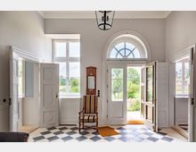 Eingangsbereich eines historischen Gebäudes mit weißen Doppeltüren, einem großen Fenster mit Rundbogen darüber und einem schwarz-weiß karierten Fliesenboden. Im Raum steht ein antiker Holzstuhl vor einem Wandspiegel, und durch die Tür ist ein Garten mit Bäumen sichtbar.