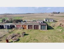 Eine Reihe von alten Garagen mit verschiedenen Holztoren steht auf einer Wiese, dahinter befinden sich ein kleines Gebäude mit Flachdach und landwirtschaftliche Felder, umgeben von Bäumen und Zäunen. Im Hintergrund sind Strommasten und eine leicht hügelige Landschaft zu sehen.